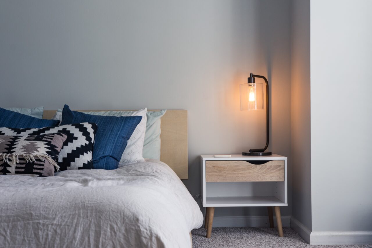 Nightstand with a light turned on next to a bed.