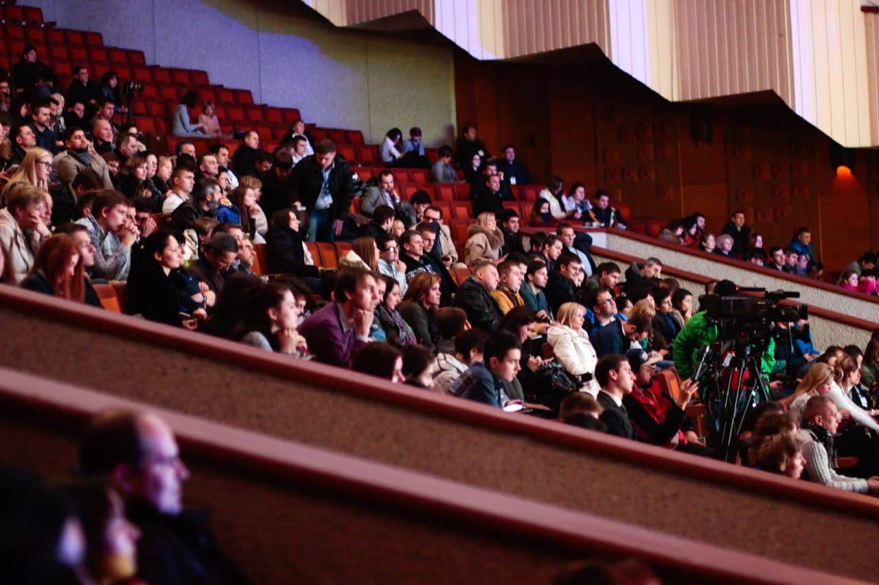 A large crowd of people watching an event from a theater balcony.