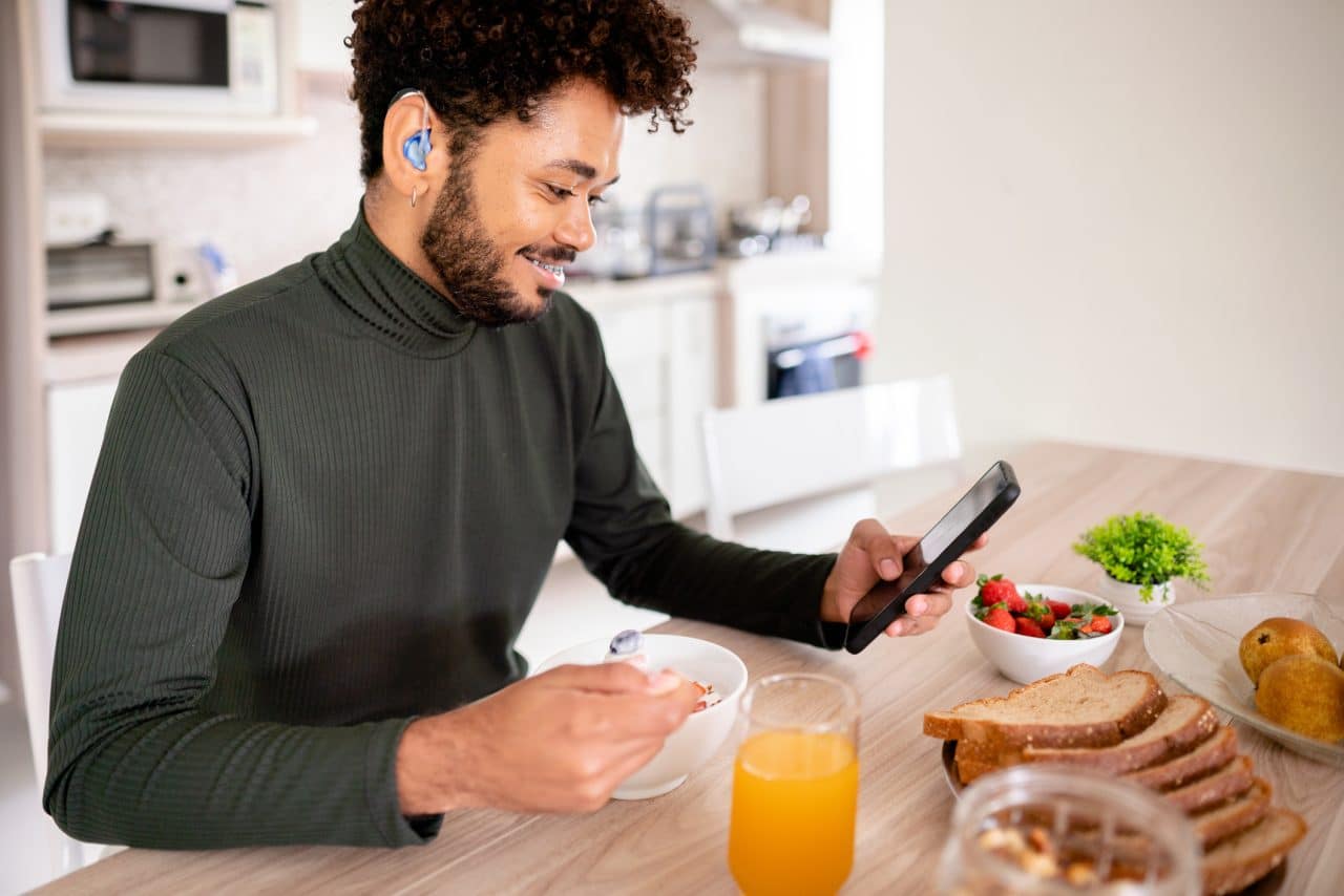 Smiling younger man with a hearing aid is checking his phone over breakfast.