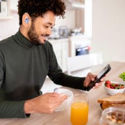 Smiling younger man with a hearing aid is checking his phone over breakfast.